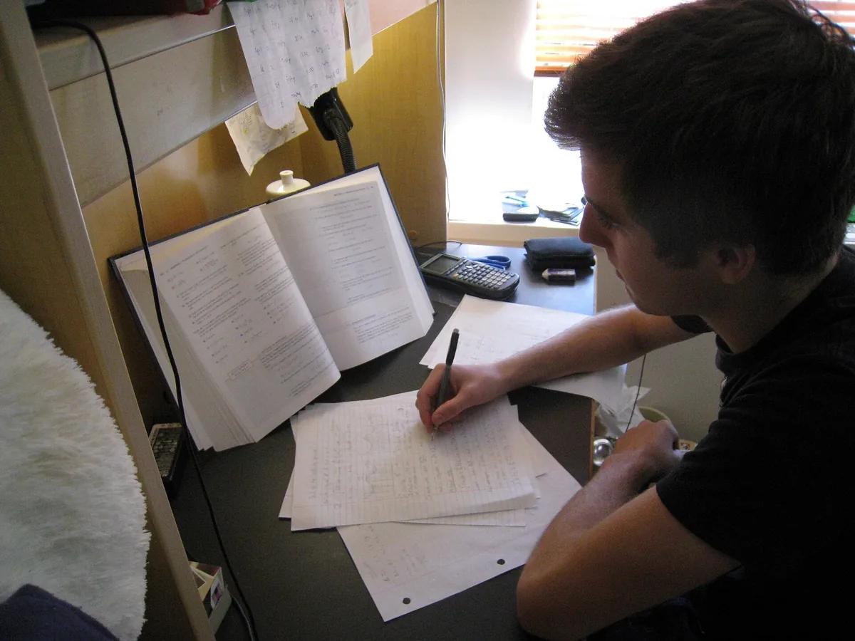 Un étudiant révise seul à son bureau, livre ouvert et calculatrice à portée de main. Photo : Gnarlycraig, domaine public