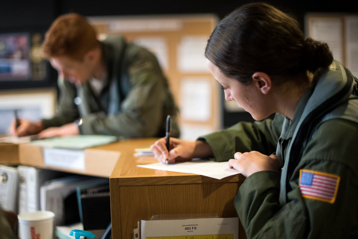 Sur une base aérienne en Angleterre, une élève officier en combinaison de vol remplit ses papiers au comptoir d'un escadron, avant un vol de familiarisation. Photo : US Air Force, domaine public
