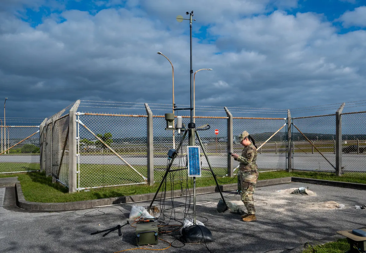 Au bord d'une piste, une prévisionniste météo relève les mesures d'un mât de capteurs, sous un ciel de cumulus. Photo : US Air Force, domaine public