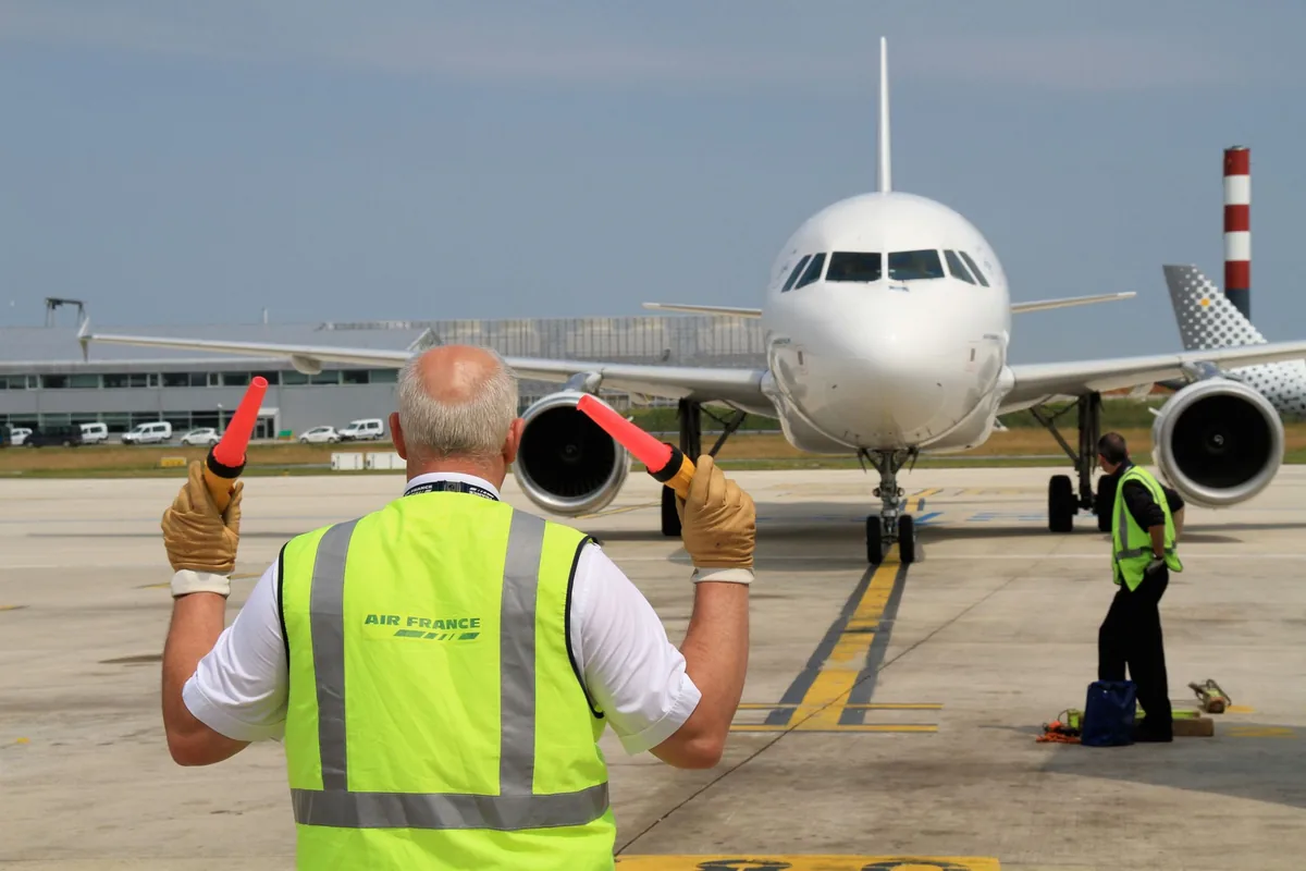 Sur l'aire de stationnement d'un aéroport, un agent de piste guide un avion de ligne jusqu'à sa place, sous l'œil d'un collègue.