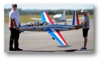 Deux modélistes portant à bout de bras une maquette volante de Fouga Magister aux couleurs de la Patrouille de France, dont l’envergure atteint environ deux mètres