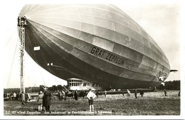 Photo ancienne en noir et blanc du dirigeable rigide Graf Zeppelin amarré à son mât près du sol, entouré d’une foule qui donne l’échelle : la coque, longue de plus de 200 mètres, porte son nom peint sur le flanc