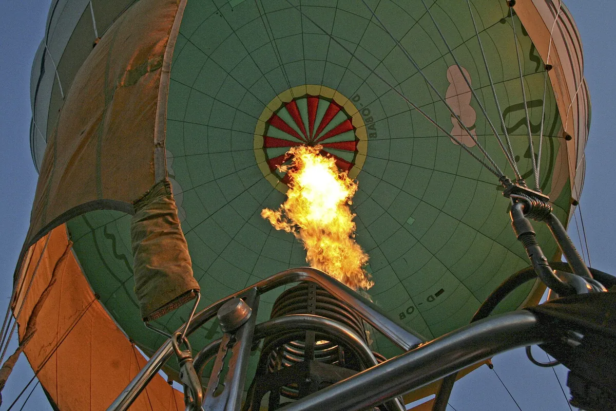 Vue depuis la nacelle d’une montgolfière, tête levée : au premier plan, le brûleur lance sa flamme dans l’enveloppe pour chauffer l’air ; tout en haut, au sommet de l’enveloppe, le disque rayé de rouge et de blanc ferme l’ouverture de la soupape, que le pilote ouvre pour laisser s’échapper l’air chaud. Photo : 5snake5, CC0