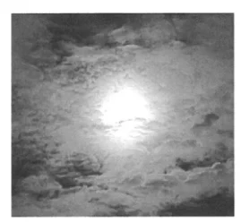 Photographie en noir et blanc du soleil, entouré d’une lueur, à travers les nuages